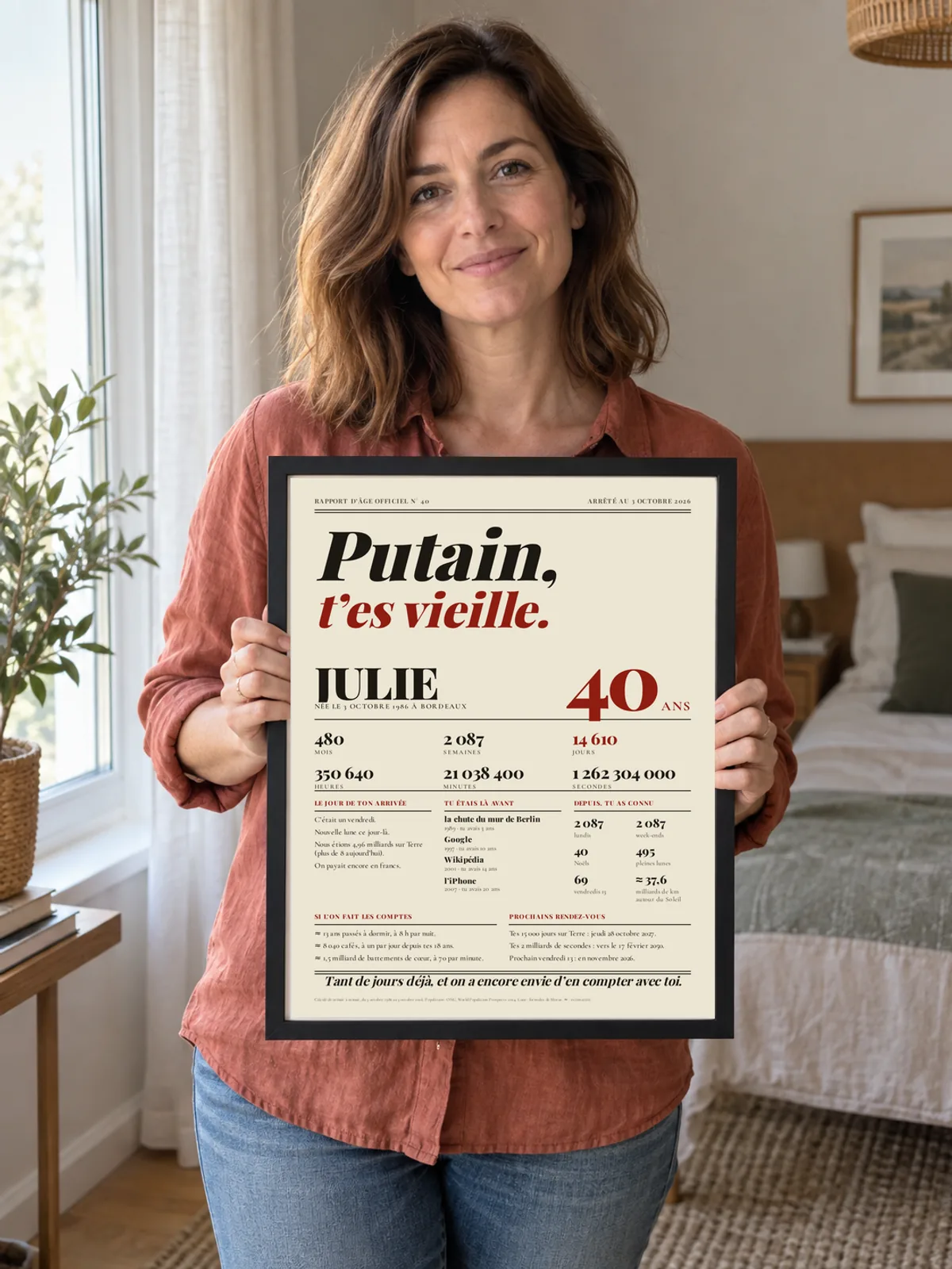 A woman in a bedroom holds up her “Bloody hell, you’re old” poster in Newspaper style, in a thin black frame