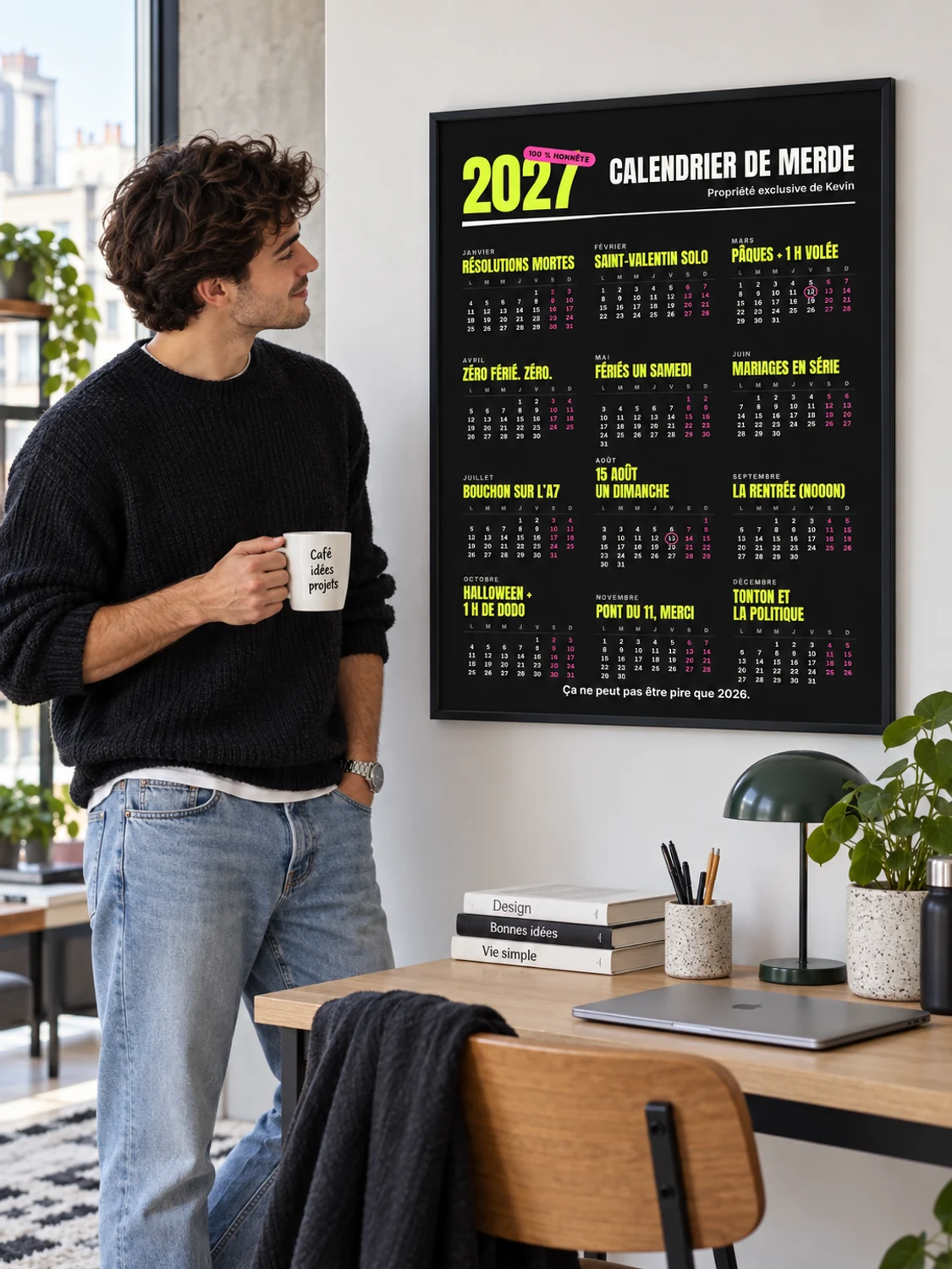 A young man holding a mug of coffee grins at a black-framed 2027 calendar poster hanging above his desk, shown here in its French version with neon yellow month names on black