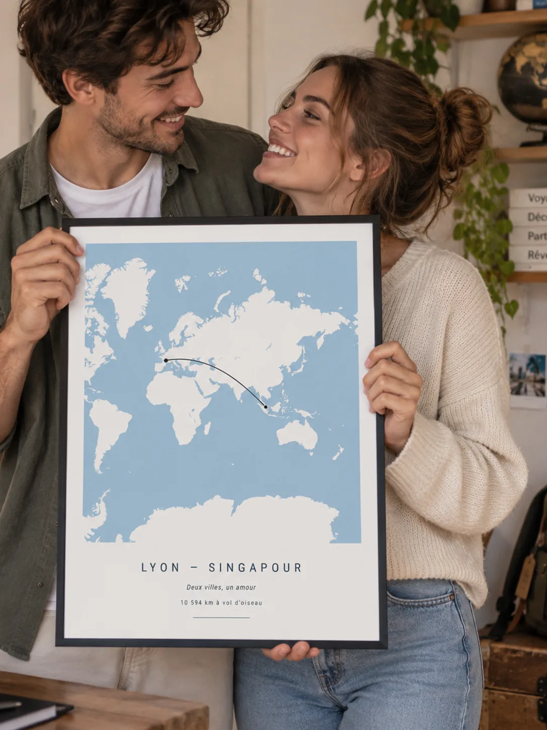 A smiling couple look at each other holding the black-framed Lyon – Singapore distance poster, “Deux villes, un amour”, 10,594 km, shown in the French version