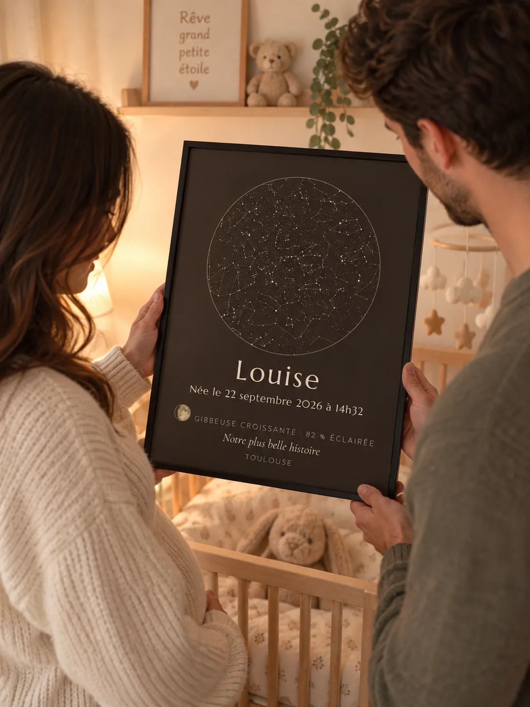 Des parents regardent l’affiche de naissance ciel étoilé de leur fille, cadre noir, dans la chambre du bébé