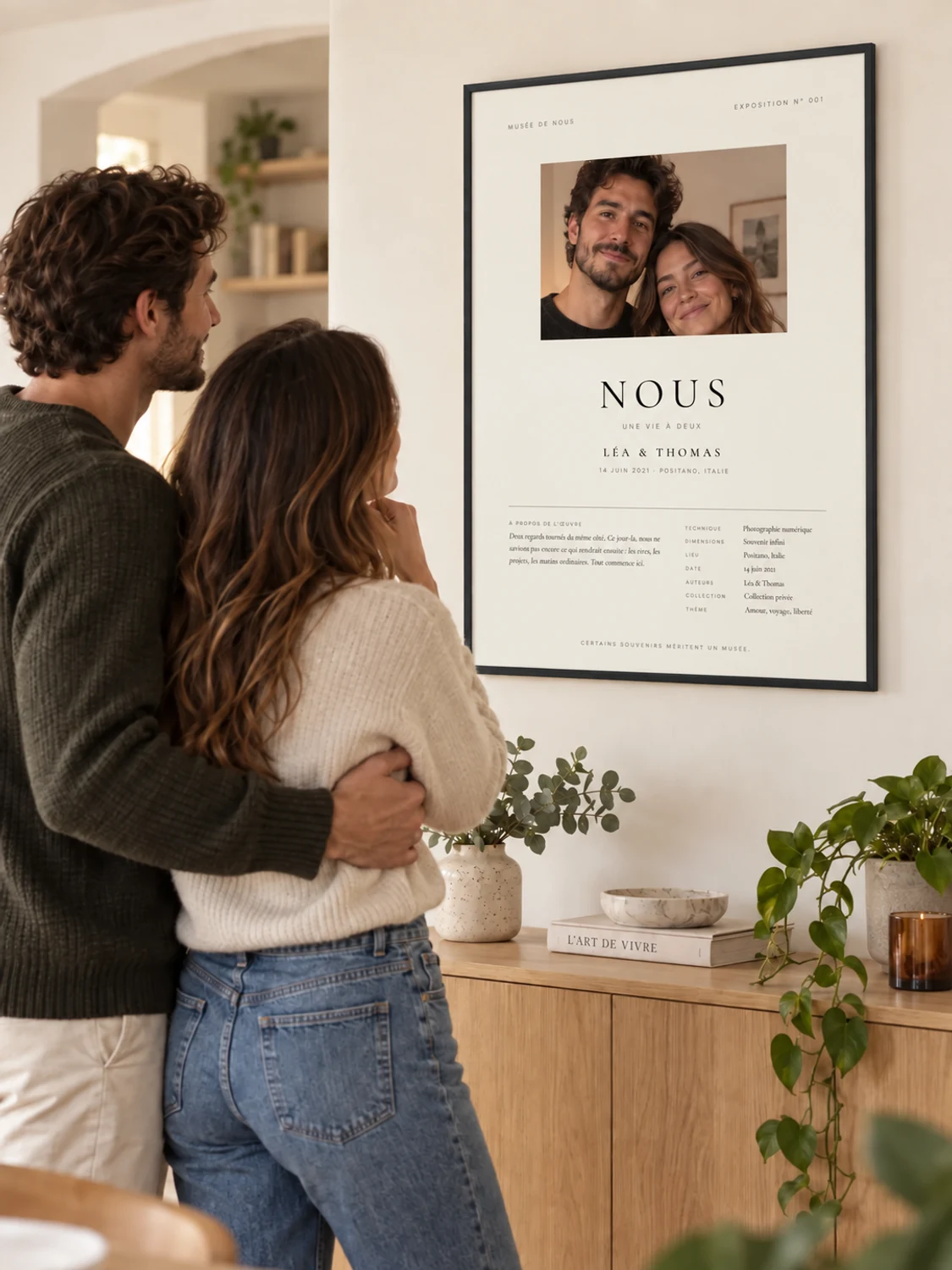 Ein Paar um die 35 betrachtet sein Foto-Poster Musée de Nous im schmalen Rahmen aus hellem Holz an der Wohnzimmerwand