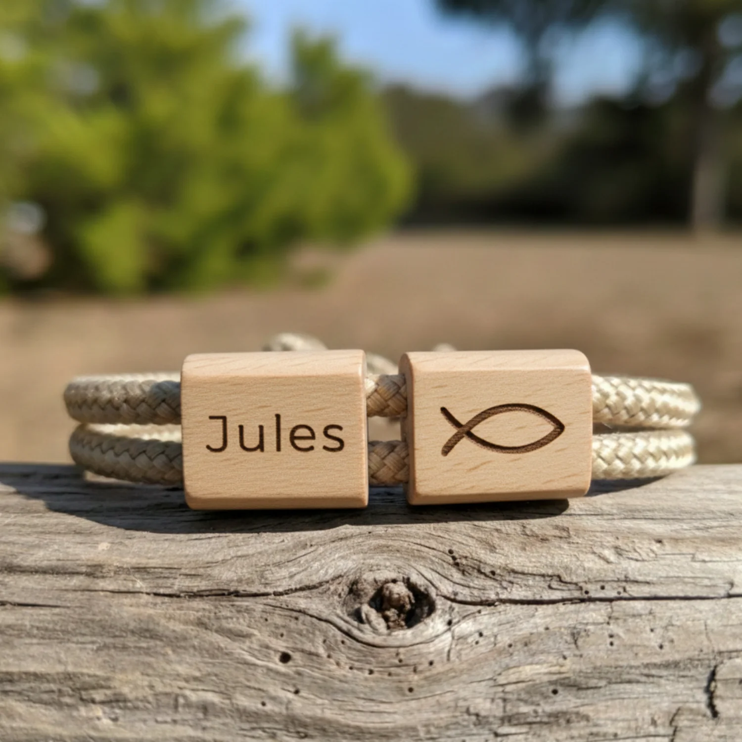 Bracelet écru gravé d'un prénom et du poisson chrétien, posé sur un bois flotté au soleil