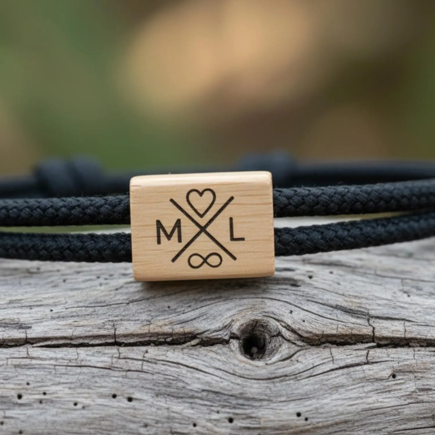 Bracelet noir gravé M et L, cœur et infini, posé sur un bois flotté gris, végétation floue en fond